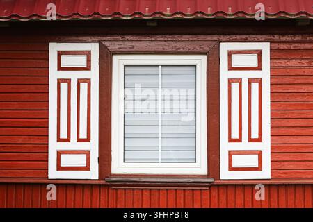 Traditionelles Fenster mit dekorativen weißen Fensterläden an einer roten Holzwand. Architekturhintergrund für die Fassade eines Bauernhauses, kulturelles Erbe und altes Design. Stockfoto