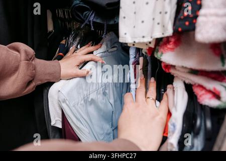 Frau durchstöbert Kleidung, die an einem Kleiderständer hängt, und wählt Kleidungsstücke aus. Modenhintergrund für die Auswahl von Garderobekleidung und das Konzept des persönlichen Stils. Stockfoto