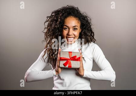 Hübsche Geburtstagfrau mit lockigem Haar und Geschenkbox auf grauem Hintergrund. Afroamerikanische Dame lächelt, sie ist glücklich mit dem Geschenk. Stockfoto
