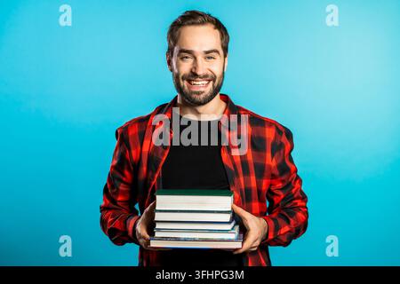 Europäischer Student in rotem kariertem Hemd auf blauem Studiohintergrund hält einen Stapel Universitätsbücher aus der Bibliothek. Der Typ lächelt, er ist Stockfoto