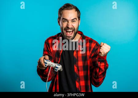 Attraktiver junger Mann spielt ein aufregendes Videospiel auf dem Fernseher mit einem Joystick an einer blauen Studiowand. Er nutzt moderne Technologie. Stockfoto