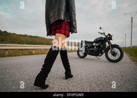 Motorradfahrerin in Minirock und Lederjacke geht zu einem retro-stylischen Motorrad. Attraktive Fahrerin in Stiefeln allein auf Stockfoto