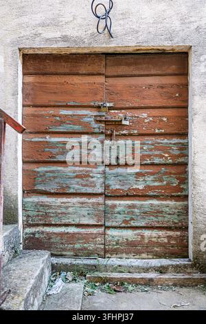 Ein altes Holztor mit abblätternder türkiser und brauner Farbe, gesichert durch rostige Vorhängeschlösser und Riegel, eingebettet in einen verwitterten Stuckrahmen mit Beton Stockfoto