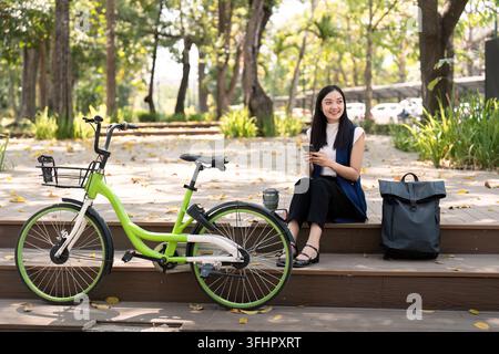 Asiatische Geschäftsfrau mit Fahrrad, die ihr Smartphone benutzt und vor dem Bürogebäude sitzt. Frau, die mit dem Fahrrad pendelt, geht zur Arbeit. Umweltfreundliches Fahrzeug, s Stockfoto