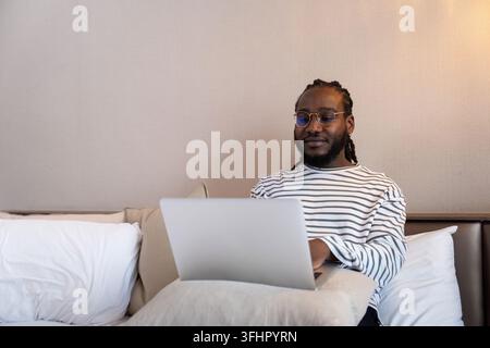 Ein lächelnder junger Afroamerikaner, der einen Laptop-Remote-Job benutzt und zu Hause auf dem Bett sitzt. Konzept der Online-Technologie-Kommunikation Stockfoto
