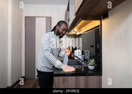 Afroamerikanischer Mann in Gummihandschuhen wischt Staub vom Tisch in der Küche, Hausarbeit, Reinigung, Lifestyle, Haushalt. Stockfoto