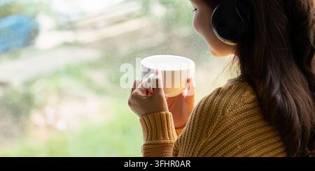 Schöne junge asiatische Frau genießt es, Kaffee zu trinken und mit Kopfhörern Musik zu hören am Schreibtisch zu Hause, Glücks- und Entspannungskonzepte. Stockfoto