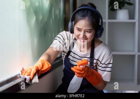 Fokussierte junge asiatische Frau, die ein Fenster mit einer Sprühflasche und einem Tuch putzt Stockfoto
