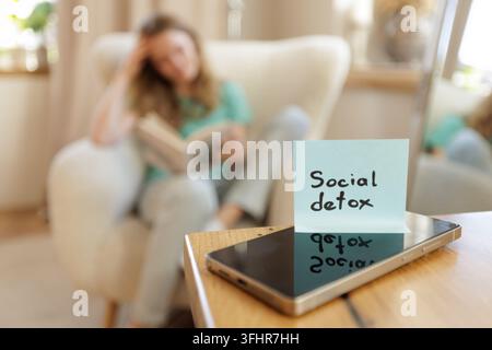 Klebezettel mit der Aufschrift „social detox“ liegt auf einem Smartphone auf einem Tisch, während eine Frau im Wohnzimmer ein Buch liest, was den Fokus auf Offline-Aktivitäten und reduzierte Nutzung fördert Stockfoto
