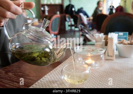 Frau, die grünen Tee aus der Teekanne aus Glas in Tassen im Café mit verschwommenem Hintergrund gießt. Hochwertige photoWoman gießt grünen Tee aus Glas-Teekanne in Stockfoto
