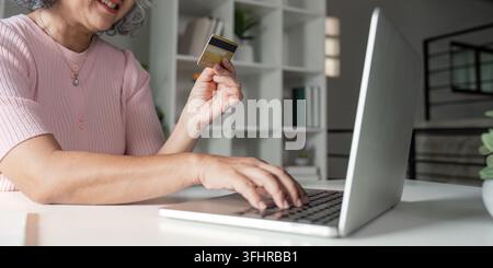 Ältere Frau, die mit Kreditkarte online einkaufen möchte, während sie in einem gemütlichen Heimbüro auf ihrem Laptop schreibt Stockfoto