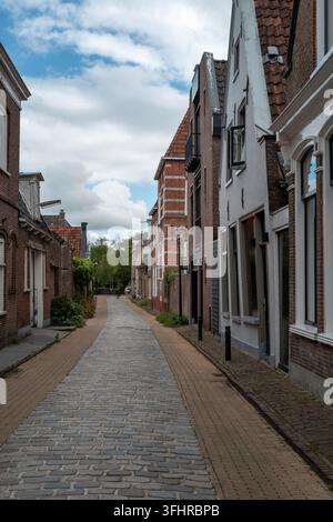 Eine schmale Straße in einer kleinen Stadt zeigt Backsteinhäuser auf beiden Seiten. Der Weg ist aus Steinen gemacht und Bäume sind im Hintergrund sichtbar. Der Himmel ist clo Stockfoto