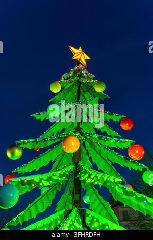Großer Weihnachtsbaum mit bunten Kugeln und hellen LED-Lichtern, der den festlichen Abend vor einem dunkelblauen Himmel erhellt und eine fröhliche Stimmung erzeugt Stockfoto