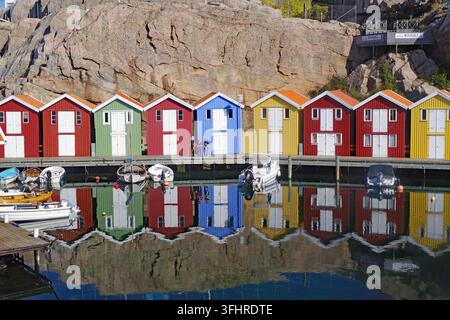Farbige Holzhäuser spiegeln sich in ruhigem Wasser, mit Felsen im Hintergrund und mehreren Booten am Pier, Smoegen, Sotenaes, Schweden Stockfoto