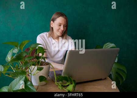 Junge Frau sitzt an einem Tisch mit Laptop zu Hause neben Zimmerpflanzen in Töpfen Stockfoto