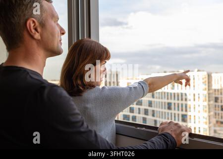 Mann umarmte seine Frau. Junges Paar zieht in neue Wohnungen. Die Leute schauen aus dem Fenster auf neue Häuser. Konzept neue Häuser, Neuanfang. Hochwertige Fotos Stockfoto