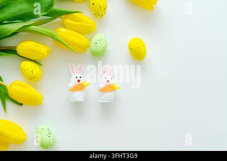 Osterdekoration mit gelben Tulpen, farbigen Eiern und zwei Hasenfiguren mit Karotten auf weißem Hintergrund. Osterfest-Konzept Stockfoto