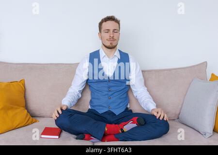Erfolgreicher junger Geschäftsmann sitzt in Lotushaltung auf dem Sofa in einem blauen Anzug Stockfoto