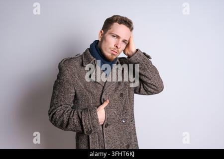 Ein junger bärtiger Mann in einem Herbstmantel posiert emotional vor einer hellen Wand. Stockfoto