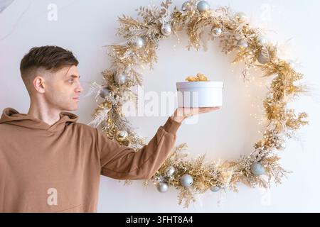 Glücklicher junger Mann hält eine Geschenkschachtel vor einem Weihnachtskranz im Hintergrund Stockfoto