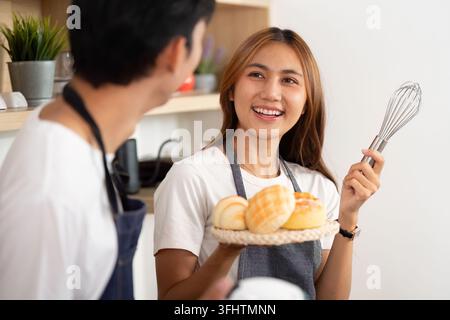 Ein fröhliches Paar teilt einen Moment in der Küche, wobei die Frau einen Teller mit frisch gebackenem Brot und einem Schneebesen hält. Stockfoto