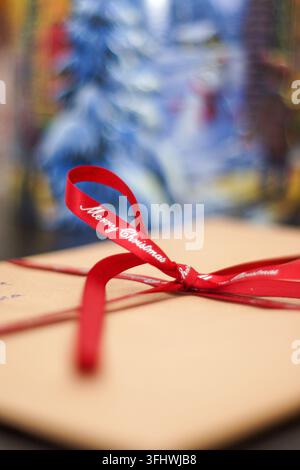 Weihnachtsgeschenk in braunes Papier eingewickelt und mit einem roten Merry-Christmas-Band verschnürt, mit einem sanft verschwommenen festlichen Hintergrund. Stockfoto