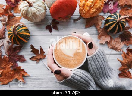 Weibliche Hände, die eine Tasse heißen Kaffee hielten, lagen im Herbst flach mit gelben Blättern und Kürbissen Stockfoto
