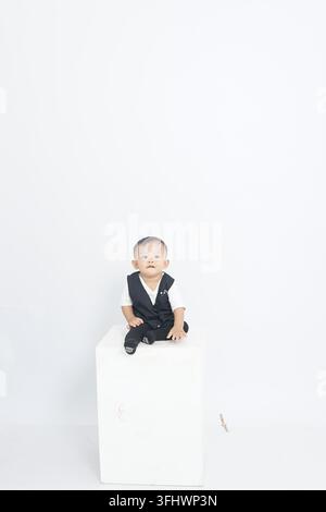 Niedlicher, liebenswerter asiatischer Babyjunge trägt eine schwarze Weste und sitzt auf einem weißen Würfel-Studio-Podest Stockfoto