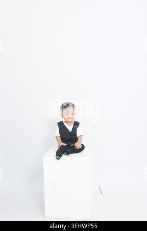 Niedlicher, liebenswerter asiatischer Babyjunge trägt eine schwarze Weste und sitzt auf einem weißen Würfel-Studio-Podest Stockfoto