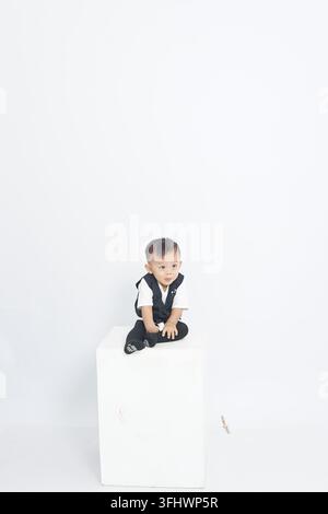 Niedlicher, liebenswerter asiatischer Babyjunge trägt eine schwarze Weste und sitzt auf einem weißen Würfel-Studio-Podest Stockfoto