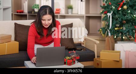 Eine Frau konzentriert sich auf ihren Laptop, beschäftigt mit Online-Weihnachts Einkäufen, umgeben von schön verpackten Geschenken. Stockfoto
