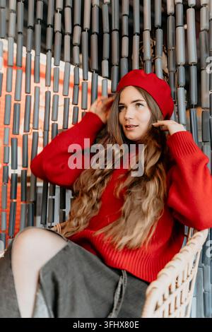 Frau mit Beret und Pullover posiert in einem Korbstuhl vor einer Perlenwand im stilvollen Café-Interieur Porträt einer bezaubernden jungen Dame Stockfoto