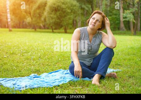 Eine erwachsene Frau sitzt auf einer Matte auf dem grünen Gras in einem Park an einem sonnigen Sommertag Stockfoto