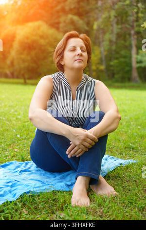 Eine erwachsene Frau sitzt auf einer Matte auf dem grünen Gras in einem Park an einem sonnigen Sommertag Stockfoto