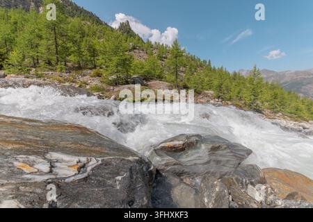 Ein rauschender Fluss kaskadiert über glatte Steine, umgeben von üppigen grünen Bäumen und majestätischen Bergen. Der hellblaue Himmel verstärkt den friedlichen Sommer A Stockfoto