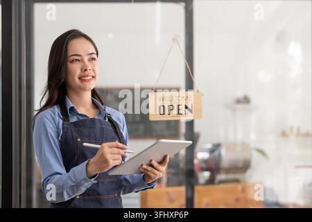 Schöne asiatische junge Barista-Frau in Schürze, die ein Tablet hält und vor der Tür eines Cafés mit geöffnetem Schild steht. Busi Stockfoto