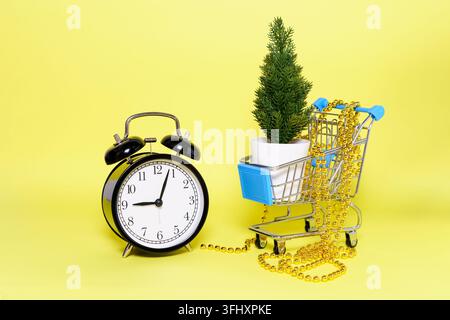 Es gibt einen künstlichen kleinen Weihnachtsbaum und goldene Perlen im Einkaufswagen, einen schwarzen Wecker auf gelbem Hintergrund. Weihnachten und Neujahr Stockfoto