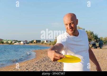 Der Mann steht am Strand und hält eine gelbe Frisbee. Er lächelt und schaut in die Kamera Stockfoto