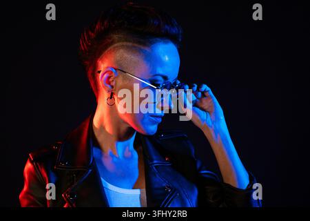 Nachtporträt einer Frau. Neonlicht. Positive Einstellung. Selbstbewusste glückliche Frau, trägt Lederjacke und Sonnenbrille, lächelt im roten und blauen Licht isoliert Stockfoto