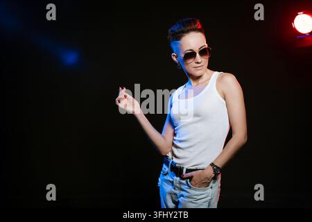 Modische Frau im Stil, posiert im Studio auf schwarzem Hintergrund. Kurzer Haarschnitt mit rasierten Seiten in Sonnenbrille und einem weißen T-Shirt. Mittelalte Kaukasierin Stockfoto