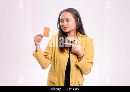 Junge südostasiatische brünette Frau vor isoliertem weißem Hintergrund, die mit einem Lächeln auf eine orangefarbene Kreditkarte zeigt und auf die Karte blickt. Stockfoto