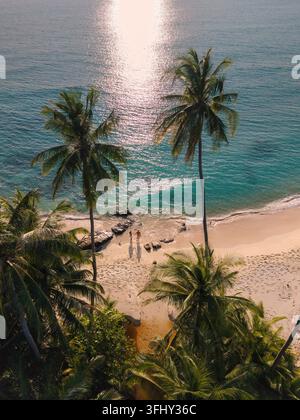 Goldenes Sonnenlicht glitzert auf den sanften Wellen, während ein Paar Hand in Hand entlang des unberührten Sandes von Takhian Beach geht, umgeben von üppigen Palmen. Stockfoto