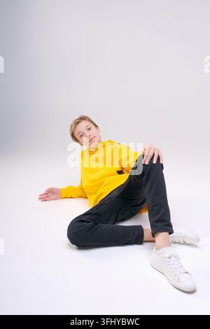 Junger, gutaussehender, glücklicher, lächelnder Teenager in schwarzen Jeans, hellgelbem Sweatshirt mit Kapuze, freut sich, während er auf einem hellen Hintergrund sitzt und posiert. Th Stockfoto