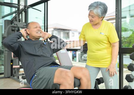 Senior-Mann, der Bauchübungen auf einem Fitnessgerät mit einer Frau macht, die ihm hilft, und repräsentiert Krafttraining und Unterstützung Stockfoto