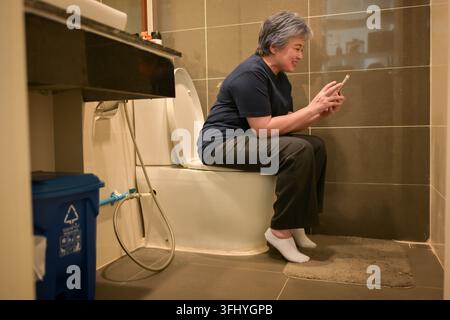 Asiatische Seniorin verwendet ein Smartphone, während sie im Badezimmer sitzt, und repräsentiert den Einsatz moderner Technologie im alltäglichen Lebensstil Stockfoto