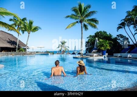 Ein Paar genießt einen entspannten Nachmittag in einem luxuriösen Pool umgeben von Palmen auf Mauritius. Das klare blaue Wasser reflektiert den hellen Himmel Stockfoto