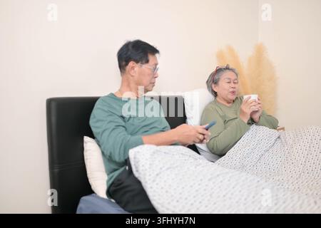 Asiatisches Seniorenpaar, das Smartphone und Tablet zusammen nutzt, während es sich im Bett entspannt und einen modernen digitalen Lebensstil zu Hause repräsentiert Stockfoto