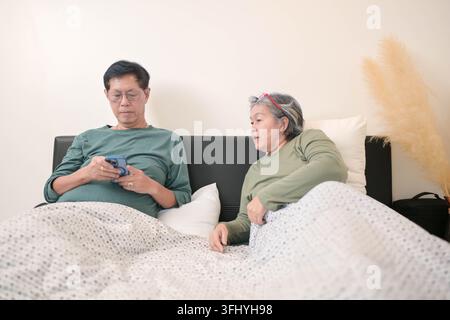 Asiatisches Seniorenpaar, das Smartphone und Tablet zusammen nutzt, während es sich im Bett entspannt und einen modernen digitalen Lebensstil zu Hause repräsentiert Stockfoto