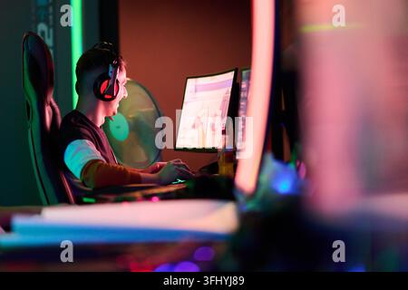 Junger Gamer, der sich intensiv auf Competitive-PC-Gameplay konzentriert, während er in einem neonbeleuchteten Raum streamt, ein Headset trägt und in einem Gaming-Stuhl neben Mo sitzt Stockfoto