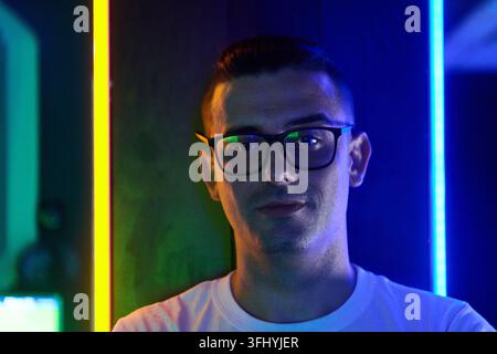 Junger Mann mit Brille, der vor vertikalen blauen und gelben Neonlichtern posiert, wodurch ein stimmungsvolles, futuristisches Urban-Porträt entsteht. Der kühle Farbkontrast ruft hervor Stockfoto